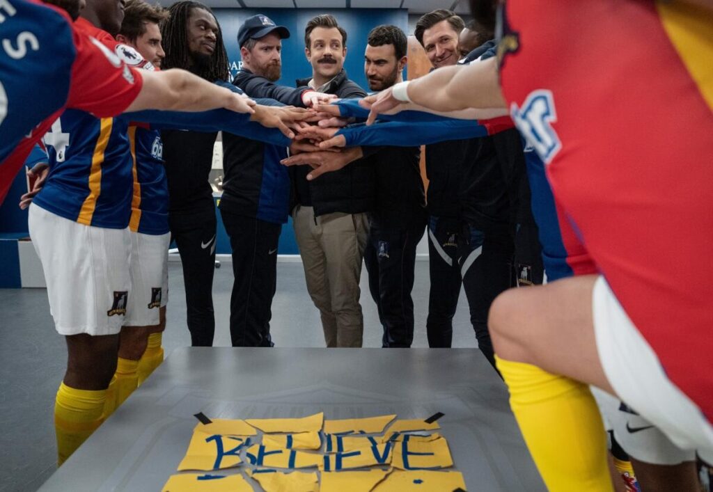 A huddle scene with a reconstructed 'Believe' sign on the foreground.